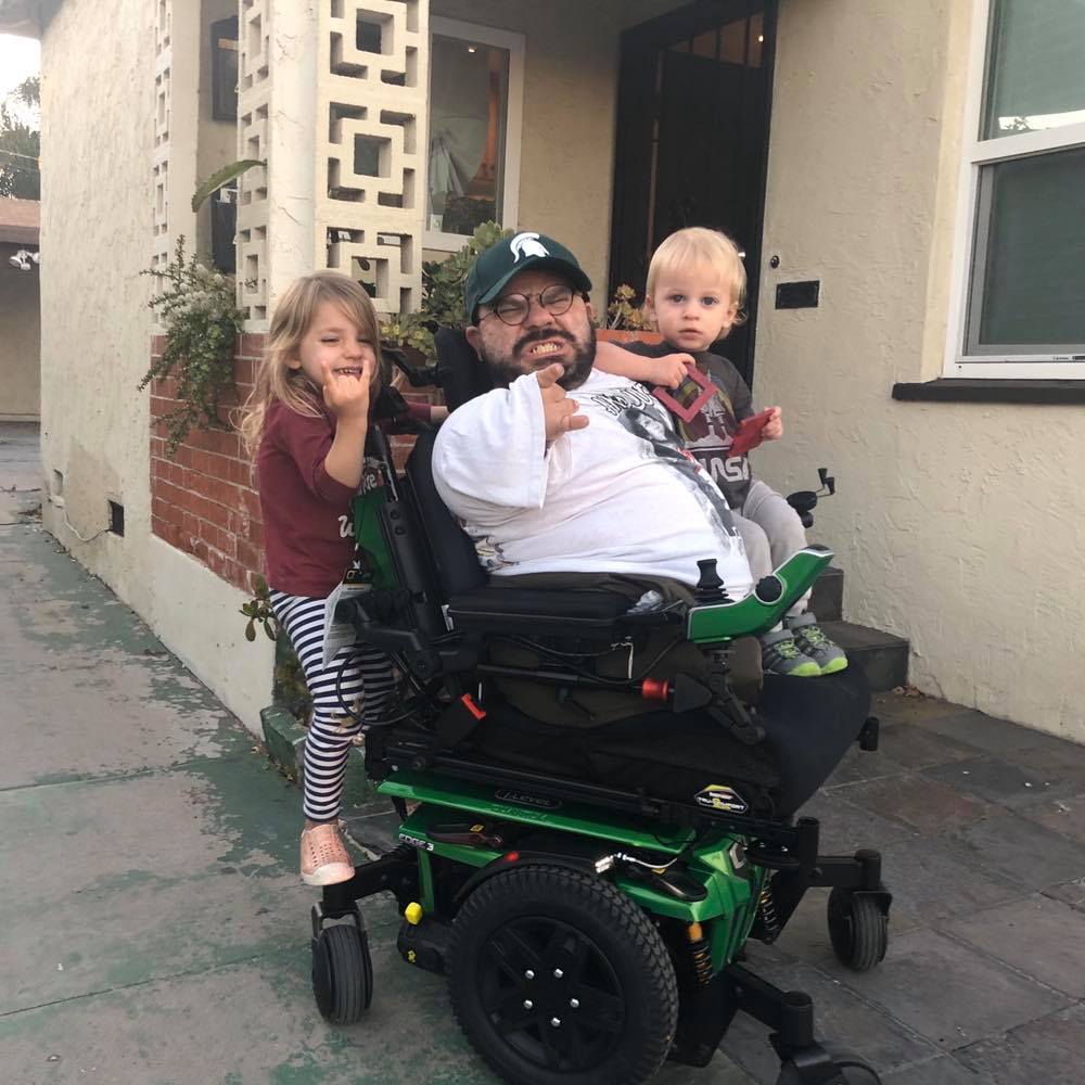 White man in a power wheelchair with two kids hanging off the back, all holding up "Rock on" signs