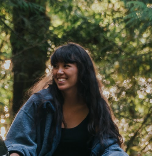 A girl with long brown hair and bangs wearing a blue jacket and black tank top smiling.