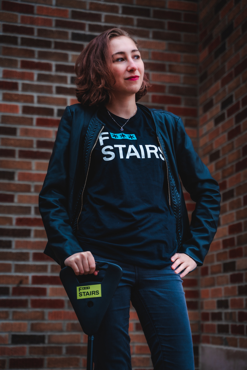 White woman with short brown hair and red lipstick wearing a f*** stairs t-shirt and black leather jacket holding her cane with a neon green f*** stairs sticker