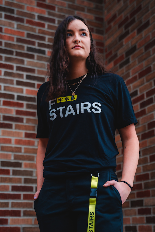 White woman with long brown hair wears a black and yellow f stairs shirt and lanyard