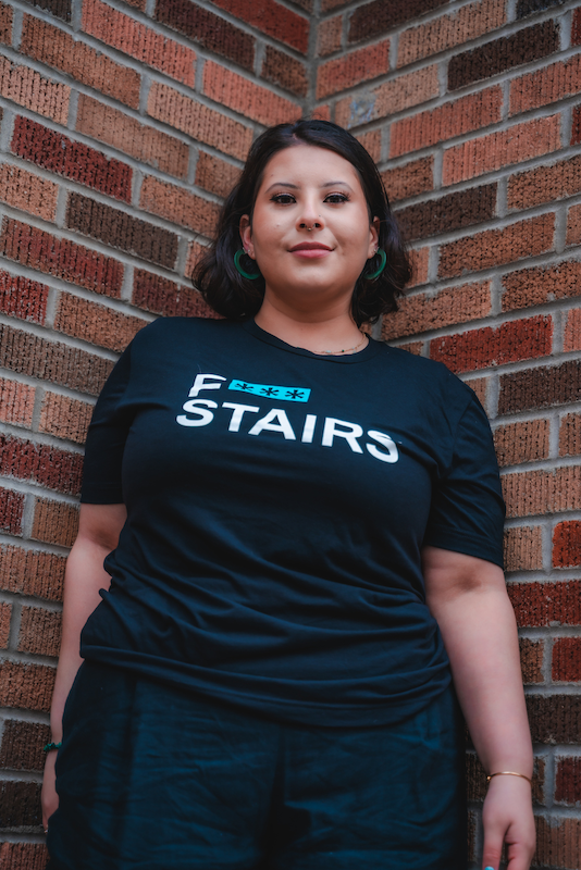 Fem presenting mixed individual with short black hair wearing a black f*** stairs t-shirt