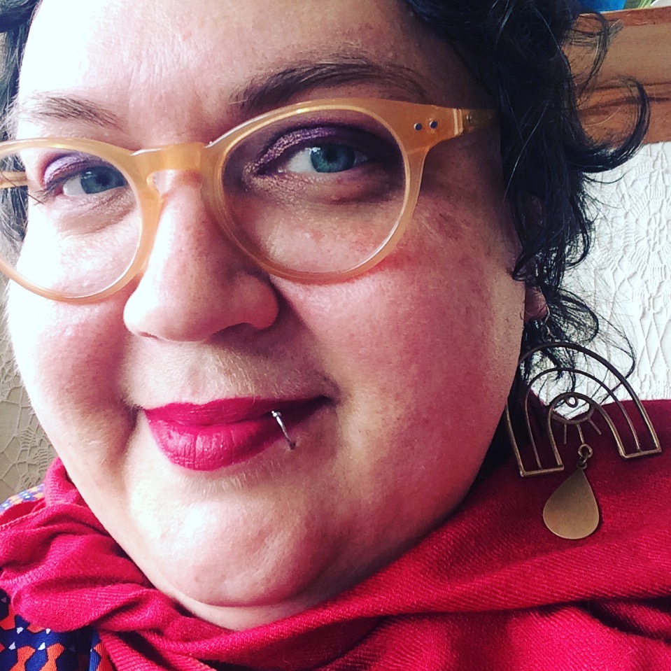 Person with glasses, red lipstick and lip piercing smiling close up to the camera.